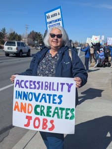Picture of former N V D A C Sectary Yvonne B. She's 5.6, grey hair, curvy, with a blue jacket and blue jeans on. She's holding a protest sign that reads, accessibility innovates and creates jobs.