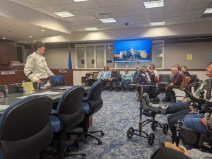 Picture is of the inside a conference room at the Nevada State Legislation Building. The room is full of people of all ages with disabilities who are preparing to make public comment to the Nevada Assemblypersons.